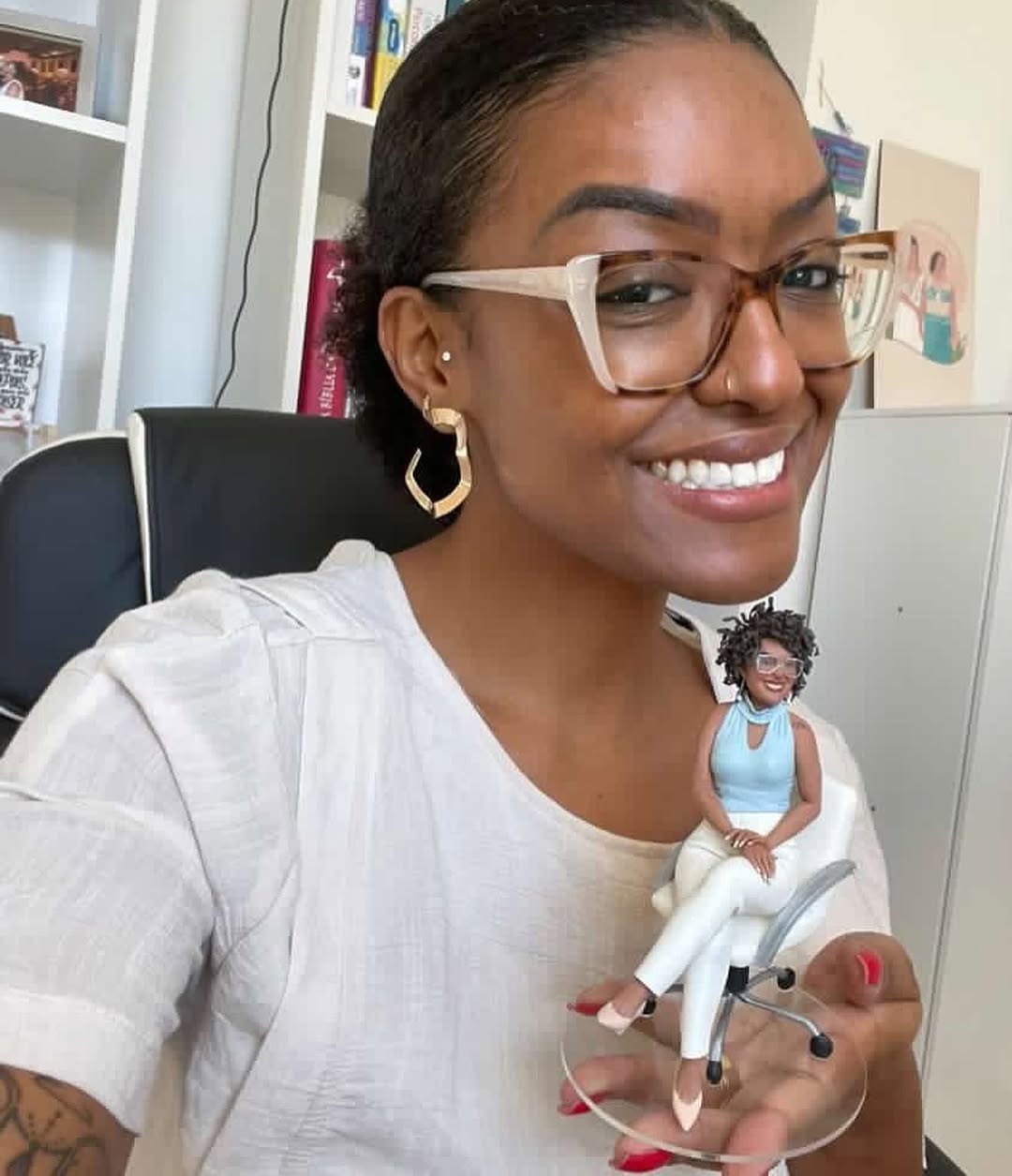 Mulher negra de óculos segurando miniatura personalizada dela mesma sentada em cadeira branca