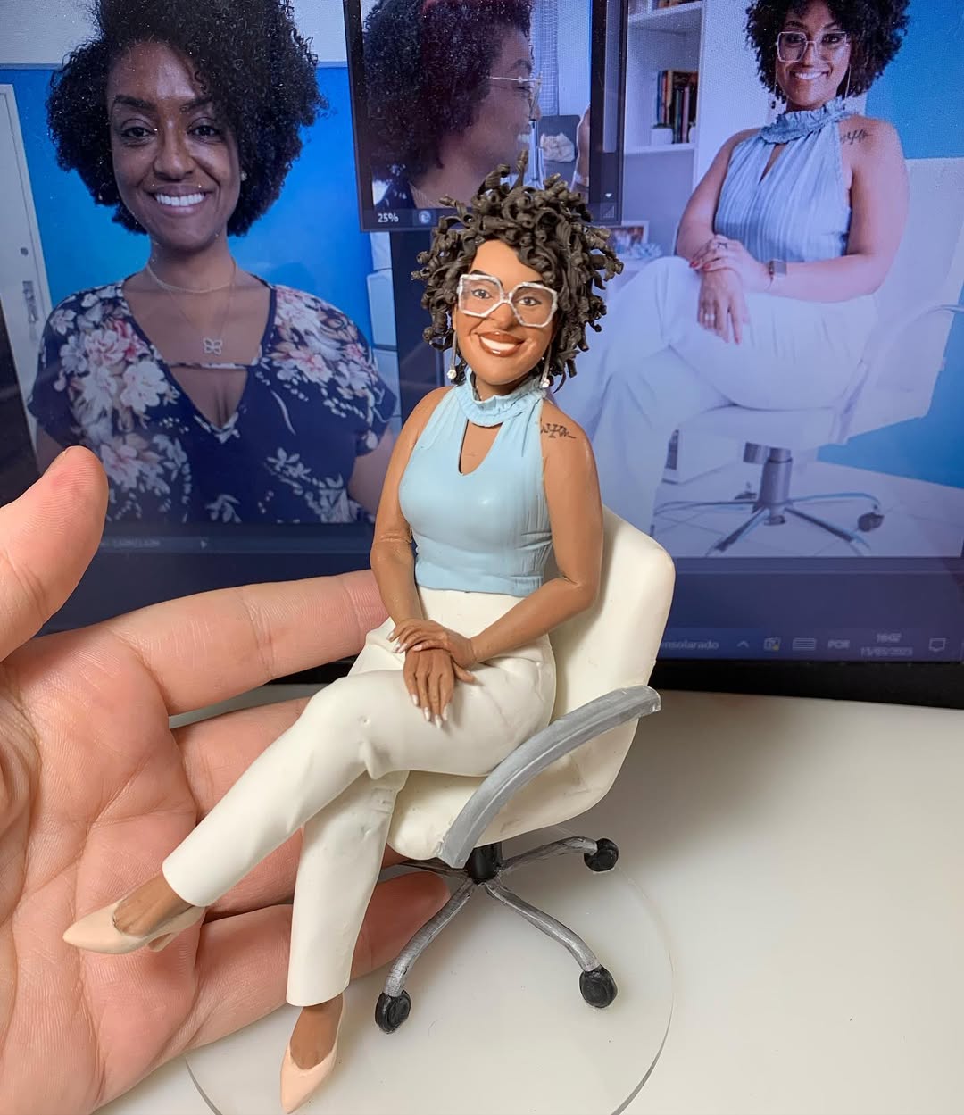 Miniatura realista de mulher negra de óculos sentada em cadeira de escritório, com fotos da pessoa real ao fundo